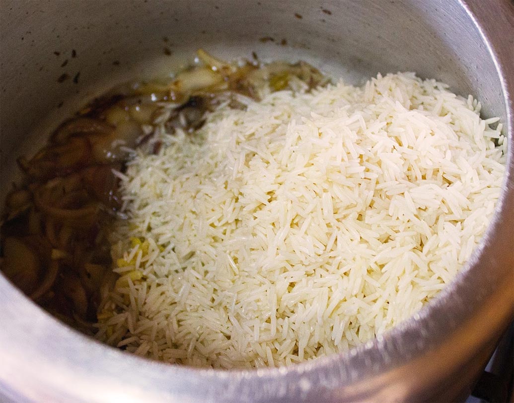 প্রেশার কুকারে ঘি গরম করে দারচিনি, ছোট এলাচ, লবঙ্গ ও তেজপাতা ফোড়ন দিন। আধ মিনিট নেড়ে নিয়ে সুন্দর গন্ধ বেরোলে বাকি পেঁয়াজ কুচি দিয়ে দিন। পেঁয়াজ সোনালি হয়ে এলে আদা-রসুন বাটা দিয়ে দিন। নাড়তে নাড়তে কাঁচা গন্ধ চলে গেলে ভেজানো চাল দিয়ে দিন।
