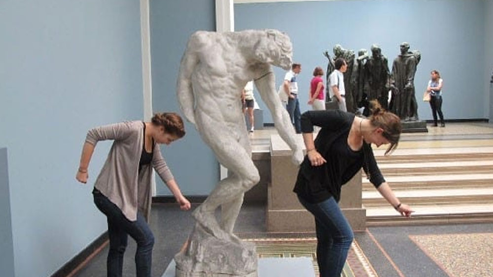 পাশের ছবিতে দেখা যাচ্ছে মিউজিয়ামে রাখা একটি সাদা পাথরের মূর্তি। হাত সোজা করে ঘাড় বেঁকিয়ে এক ব্যক্তি দাঁড়িয়ে আছেন, এই ভঙ্গিতে মূর্তিটি বানানো হয়েছে। মিউজিয়াম ঘুরতে গিয়েই দুই মহিলার চোখে পড়ে মূর্তিটি। তাঁরা মূর্তির ধাঁচ নকল করে এমন ভাবে ছবি তোলেন, যেন এটি নাচের কোনও বিশেষ ভঙ্গিমা।