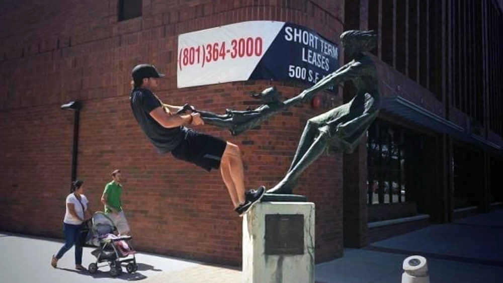 পাশের ছবিতে পাথরের মূর্তিটি লক্ষ করলে দেখা যায়, এক মহিলা একটি বাচ্চা মেয়ের হাত ধরে ঘোরাচ্ছেন। কালো টি-শার্ট পরা ব্যক্তিটি সেই পাথরের মূর্তির ধারে এমন ভাবে উঠে দাঁড়িয়েছেন যে দেখলে মনে হবে, তিনি এবং মূর্তির মহিলাটি দু’দিক থেকে বাচ্চাটির হাত-পা ধরে টানাটানি করছেন।