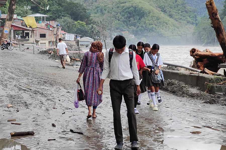 নদীর জল নেমে যাওয়ার পর বহু রাস্তার উপর জমে রয়েছে পলিমাটি। তার উপর দিয়েই স্কুলে যাচ্ছে পড়ুয়ারা। রাস্তায় দাঁড়িয়ে থাকা গাড়ি, দু’চাকা আটকে রয়েছে কাদামাটিতে। 