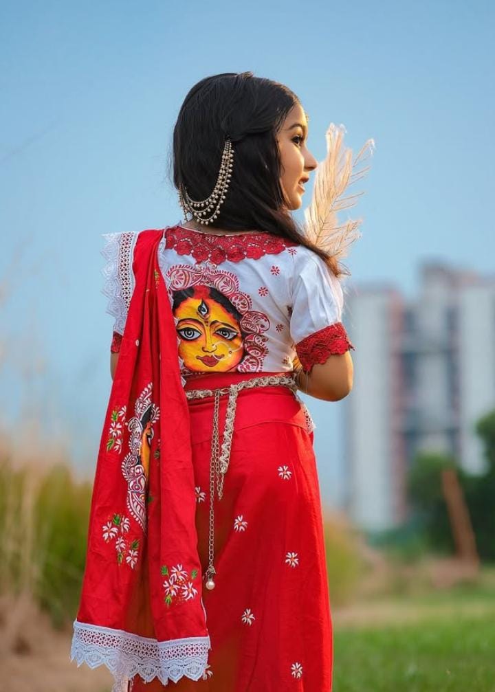 তার ব্লাউজের পিছনে আঁকা দেবীর মুখ– হলুদ রঙের মুখশ্রী, বড় বড় চোখ, লাল টিপ। যেন শরতের মাঠে কাশের বনের ফাঁকেই উঁকি মারছে দেবীর প্রতিচ্ছবি।