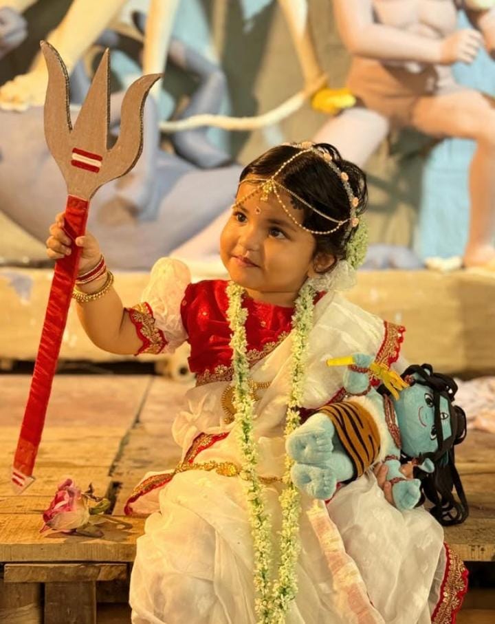  লাল-সাদা পোশাকের সঙ্গে চুলে জড়ানো জুঁইফুলের মালা, কপালে টিকলি, গলায় ভারী হার। কোমরে কোমরবন্ধ। হাতে ধরা পদ্মফুল। একেবারে সাক্ষাৎ খুদে লক্ষ্মী যেন! 