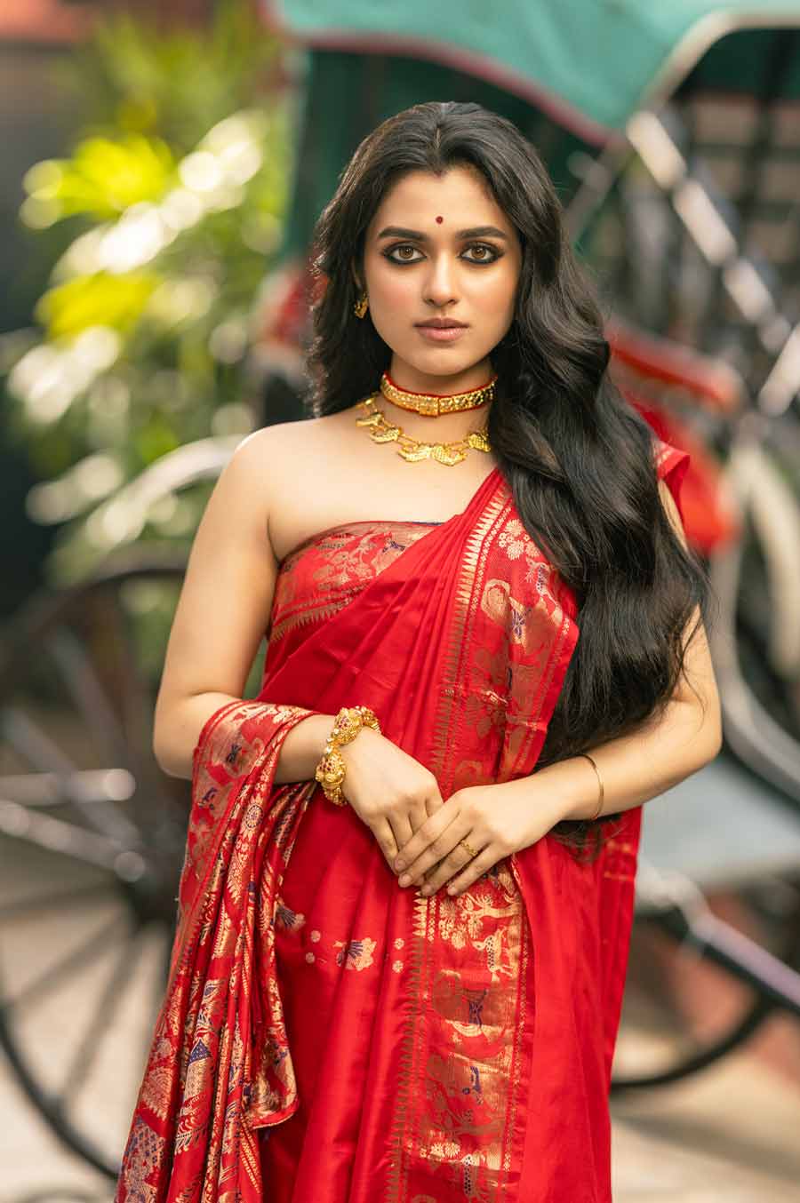 শ্যামৌপ্তিকে সাবেকি ধাঁচে শাড়ি পরানো হয়েছে এখানে। ব্লাউজ় ছাড়া শাড়ি। আঁচলের কায়দায় রয়েছে বৈচিত্র। আটপৌরে ভাবে পরানো লাল টুকটুকে স্বর্ণচরি শাড়ির আঁচল কখনও কাঁধে ফেলা, কখনও বা হাতে। এক ঢাল কালো চুল ফেলে রাখা হয়েছে ঘাড়ে। ছোট বোনকে ঘিরে অনুপমের শৈলীর ছাপ চোখে পড়ার মতো। তাঁর কথায়, ‘‘টিউব টপ, ব্রা, ট্যাঙ্ক টপ ইত্যাদিকে ব্লাউজ় হিসেবে পরার চল বেড়েছে আজকাল। ফলে এখন নানা ভাবে সাবেক সাজকে ফিরিয়ে আনা হচ্ছে। তার উপর গয়নাগুলোও একটু অন্য রকমের।’’ শ্যামৌপ্তির গলায় মাছ মোটিফের হার এবং ঠোকাইয়ের চোকার, হাতে বাঘ বালা, আঙুলে আংটি। সোনার গয়নায় মুড়ে, সাবেক সাজে ঝলমল করছেন শ্যামৌপ্তি।