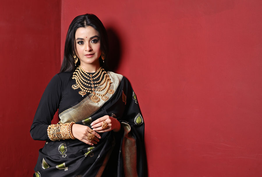 বৌভাতের সাজে সম্পূর্ণ ছকভাঙা সাজে সেজেছেন দর্শনা। কনে মানেই যেখানে উজ্জ্বল রং, সেখানে হাতে বোনা কালো বেনারসি পরেছেন অভিনেত্রী। শাড়ি জুড়ে ফ্লুরোসেন্ট সবুজের মিনাকারির কাজ। ব্লাউজ়ের বদলে কালো ফুল-হাতা বডিস্যুট পরানো হয়েছে তাঁকে। যাতে দেহের আকার সুন্দর দেখায়। দর্শনা এখানে লাল ছোট্ট টিপ পরেছেন। আপনি চাইলে কালো বা মেরুন টিপ দিয়েও সাজ সম্পূর্ণ করতে পারেন।