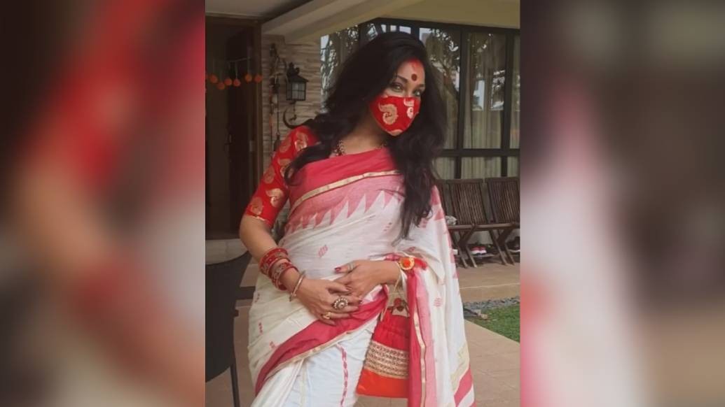 লাল পাড় সাদা শাড়ি, বড় লাল টিপের চিরন্তনী সাজে অপরূপা ঋতুপর্ণা সেনগুপ্ত। লাল বেনারসী ব্লাউজের সঙ্গে ম্যাচ করা মাস্কে তাল মেলাল করোনা-সময়।