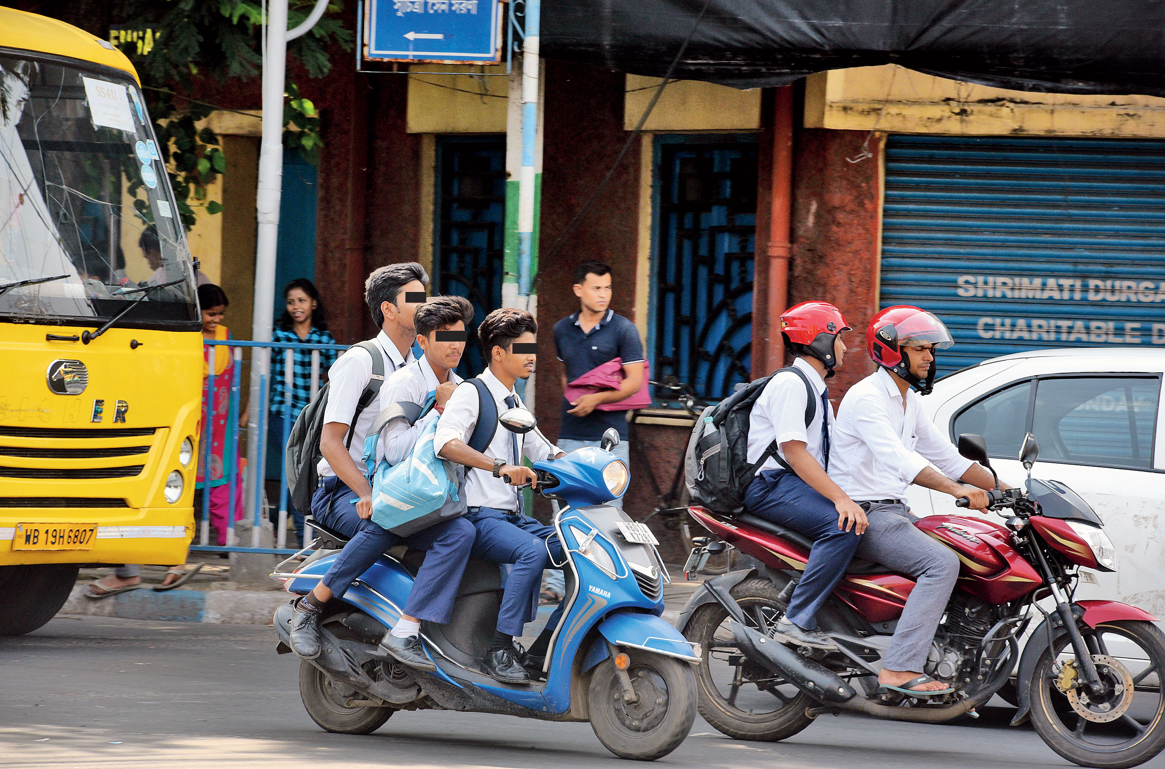 Three boys in school uniform ride a two-wheeler without helmets near Ballygunge Dhaba.