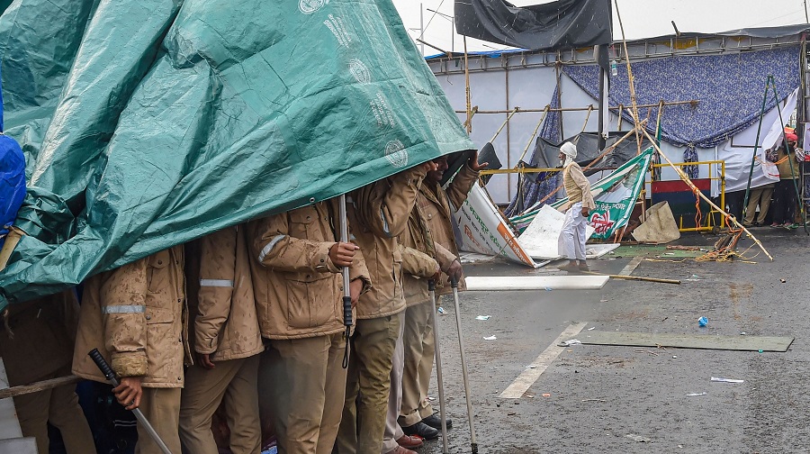 Police personnel take shelter under a waterproof tarpaulin sheet during rain near the farmers protest site, at Delhi-UP border near Ghazipur, on Tuesday.