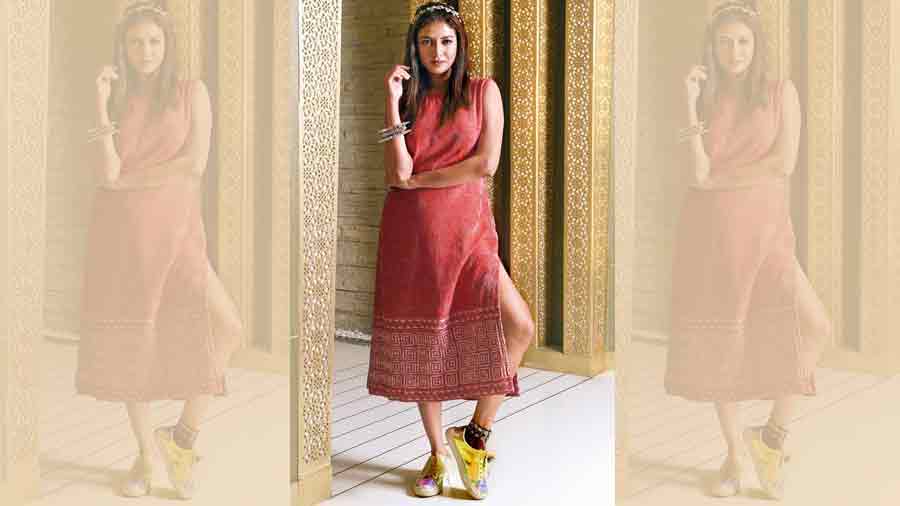Sauraseni channels an effortless Shasthi look in this laal-shada shift dress from SHE Kantha designed with kantha embroidery on crepe. A perfect blend of the traditional and the contemporary, the inverted use of laal-paar-shada and kantha is designed in a contemporary style with an optical illusion design on the fabric. Fresh and nude face, hair worn straight and accessorised with a necklace used as a headband, chunky bangles, a bracelet used as an anklet and comfy sneakers, complete the look.