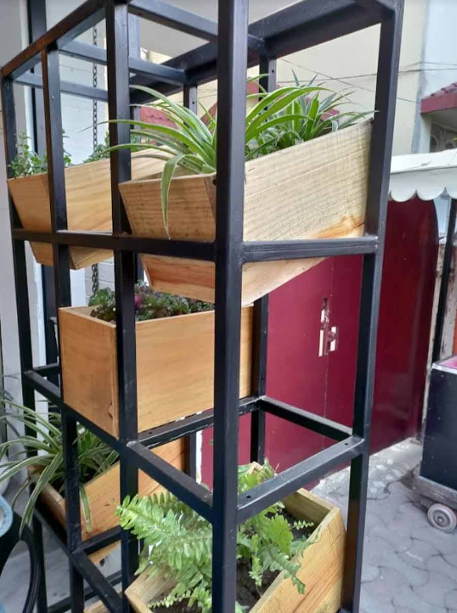 The outdoor seating patio section has a stylish wrought-iron shelf that has been used to hang plants from wooden boxes. It adds a subtle freshness and livens up the dark furnishing with a smattering of green