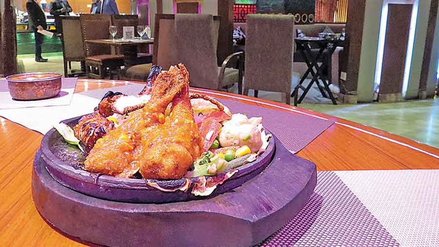 Tandoori Jhinga and Prawn Nuggets served on a sizzler with the glass and teakwood decor of the seating area of Orko’ss in the backdrop.  (Top) Barbecued chicken winglets.