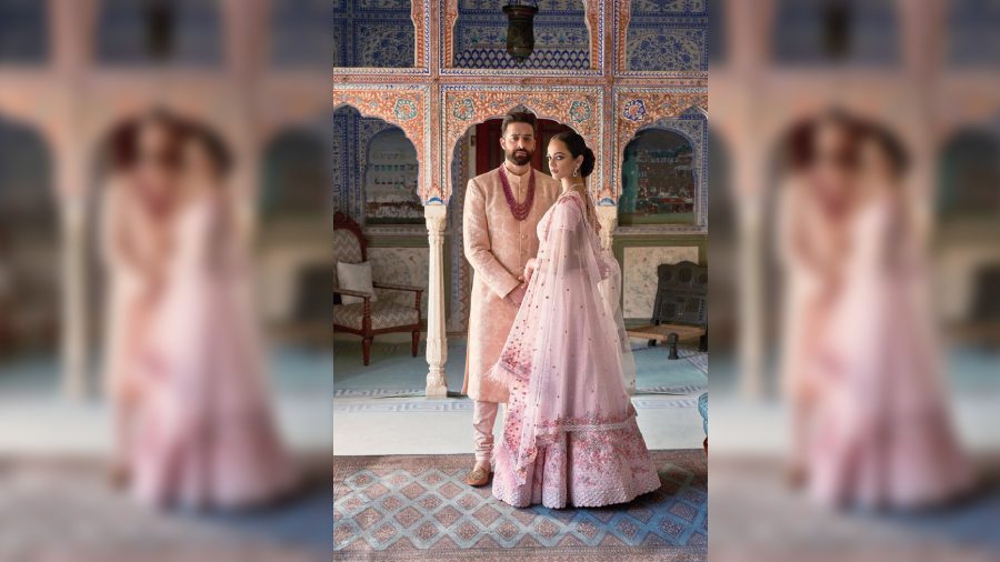 His buff pink kantha achkan + matching kurta + churidar complements her blush pink silk lehnga + matching blouse + feather-trimmed tulle dupatta draped like a cape. Crystals, beads and silk thread embellishes the lehnga.
