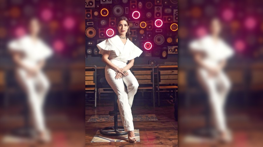 A subtle-glam, chic western party look in all-white for summers, Richa posed against the glow light speaker-design wall in an exaggerated sleeve top and broad pant ensemble from Sixth Avenue, accessorised with a sleek diamond and emerald neckpiece and finger ring, bright lips and open hair.