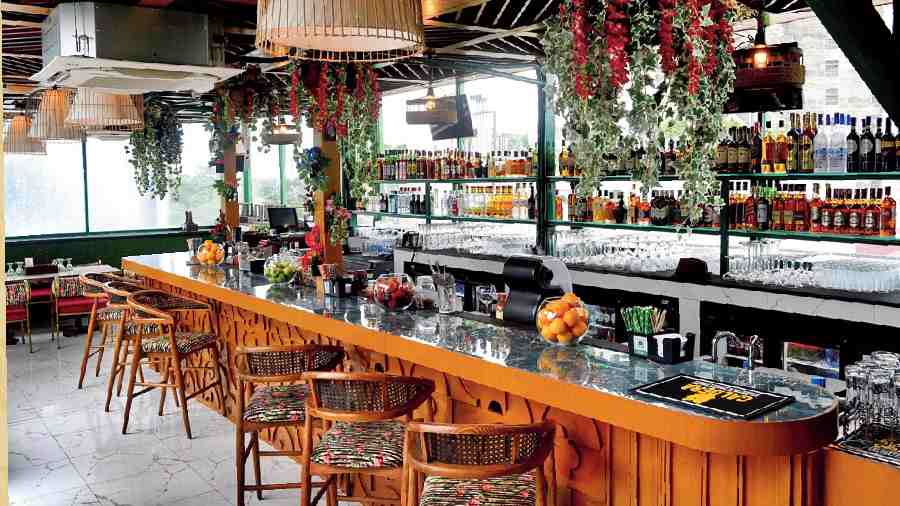 The bar in the glasshouse has a cute European vibe with plants and flowers hanging from the top. It is done in woody shades with bright printed velvet high chairs. Cane lamps line the ceiling, and the rest of the velvet seating theme follows throughout the glasshouse.