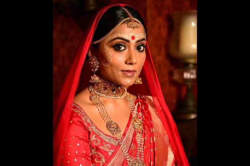 Keeping the Bengali bridal look traditional to the T, Abhijit draped a red handwoven golden and silver alfi weave Benarasi on Anindita. The low middle-parted bun and simple make-up were completed with black kohl eyes, giving a brown smokey touch to it, and semi-nude lips. The big red bindi on the forehead added the traditional and authentic Bengali bridal touch to the look, highlighted with the bhorat chandan style. The kalka motifs extend from the forehead to the cheek and chin has dainty designs in this style.