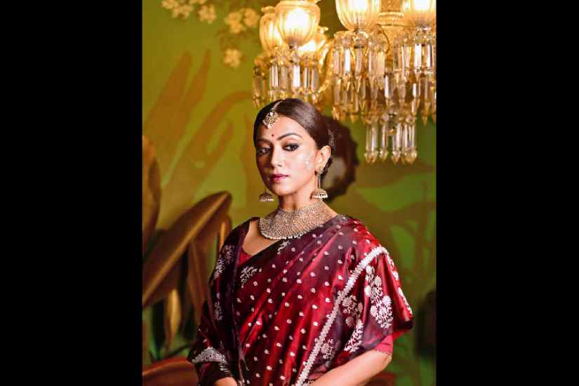 For brides who want to ditch the bright red and go for minimal make-up with a traditional touch, this look is apt for the D-day. Anindita wore a handwoven silver kadhua Benarasi with jamdani weave border. The dark maroon sari is complemented with simple make-up with classic kohl eyes, a small bindi and middle-parted bun. Kaan chandan style of chandan art highlights this look with a combination of kalka and dot motifs drawn beside the ears. Perfect for those who don’t want to keep the chandan art in an elaborate manner