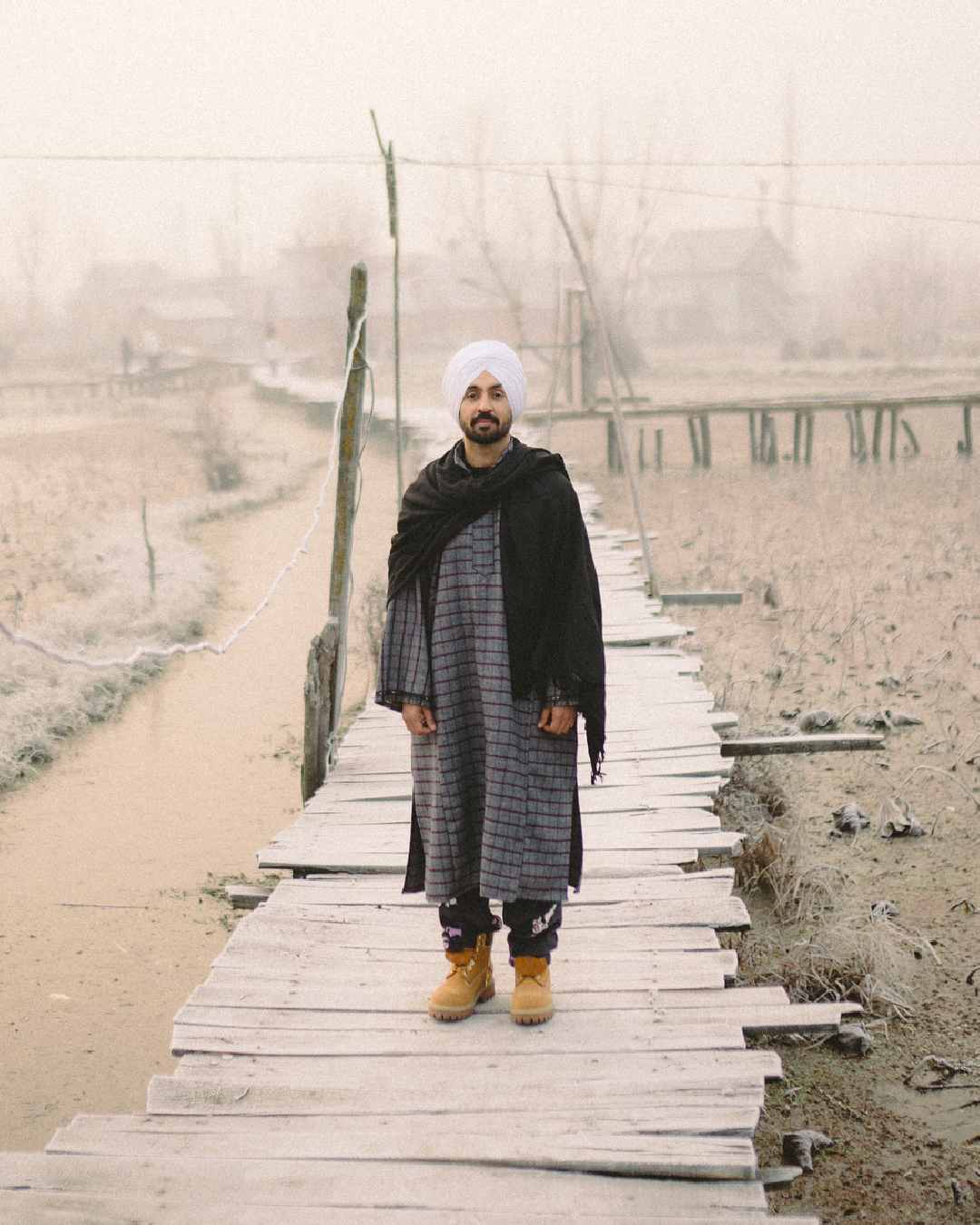 Dressed in a traditional pheran with a black shawl wrapped around his shoulders, Diljit Dosanjh posed for a picture on a wooden bridge in Kashmir.