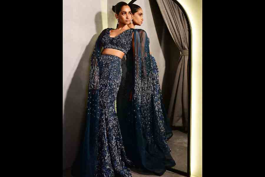 Meghna poses elegant in a deep sea blue mermaid lehnga, paired with a chic blouse adorned with sequins, beads and pearl work. The embroidered sheer trail cape adds a trendy touch, making it perfect for a sangeet evening.
