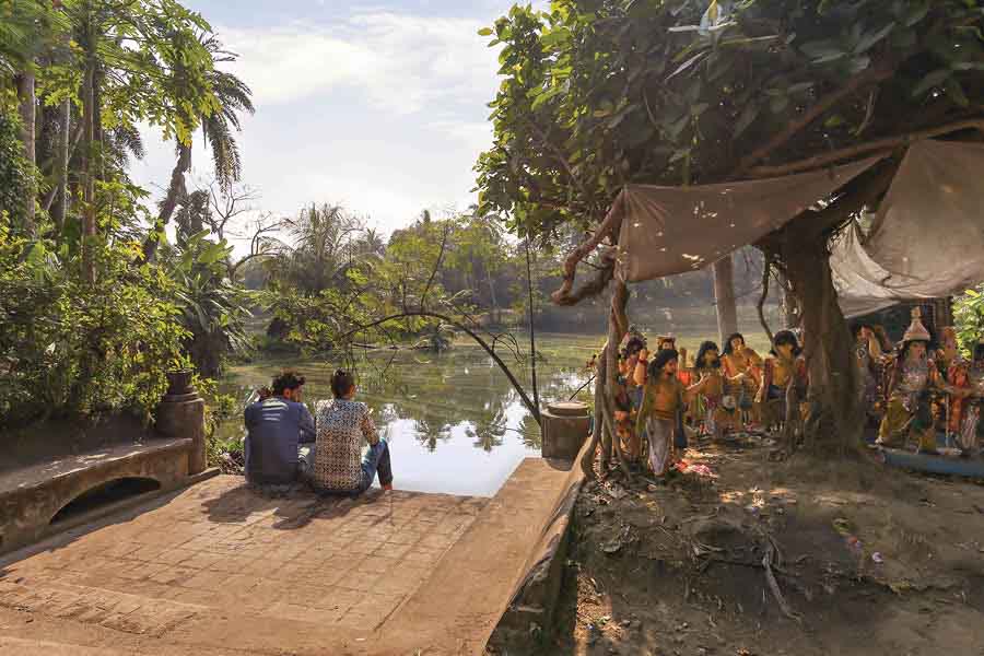 Friends sit and gossip beside a village pond with rural landscape at Deulti, West Bengal