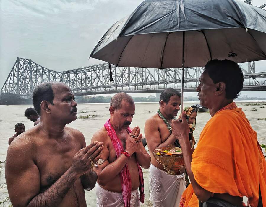 Beneath the Howrah Bridge, this priest holds on to an umbrella amid the rain even as he guides devotees to utter mantras for their ancestors