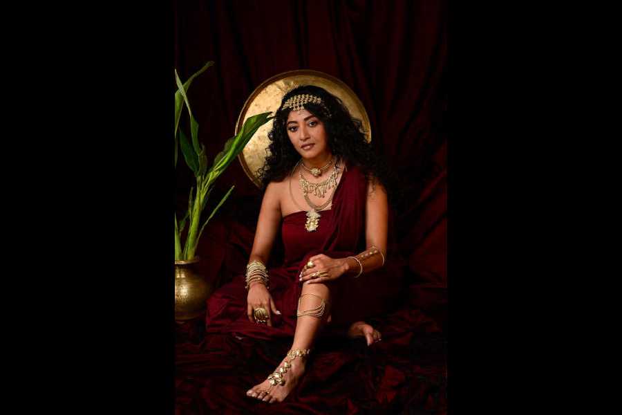 Paoli exudes elegance in a maroon sari that beautifully complements her look in metallic shades. A mathapatti featuring dainty rose motifs and layered neckpieces that includes a striking choker with a lion crest, a necklace themed on teardrops merging, and a pendant depicting Durga, accessorise her look. Her hands are adorned with a chic cylindrical cuff and a playful spring cuff, both designed with matching rose motifs, along with finger rings featuring lotus, soldier, and stone motifs. A foot piece inspired by the gentle curves of rose petals adds a finishing touch to the look. Shimmery hues of gold on her chest and hands and copper hue enhancing her eyes and lip complement the hair worn in open curls.