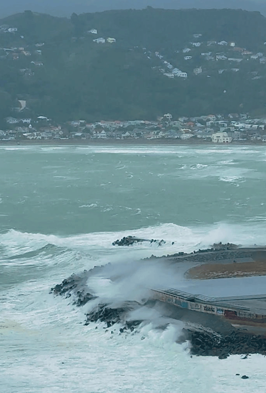 A large wave breaks over the breakwater at Lyall Bay