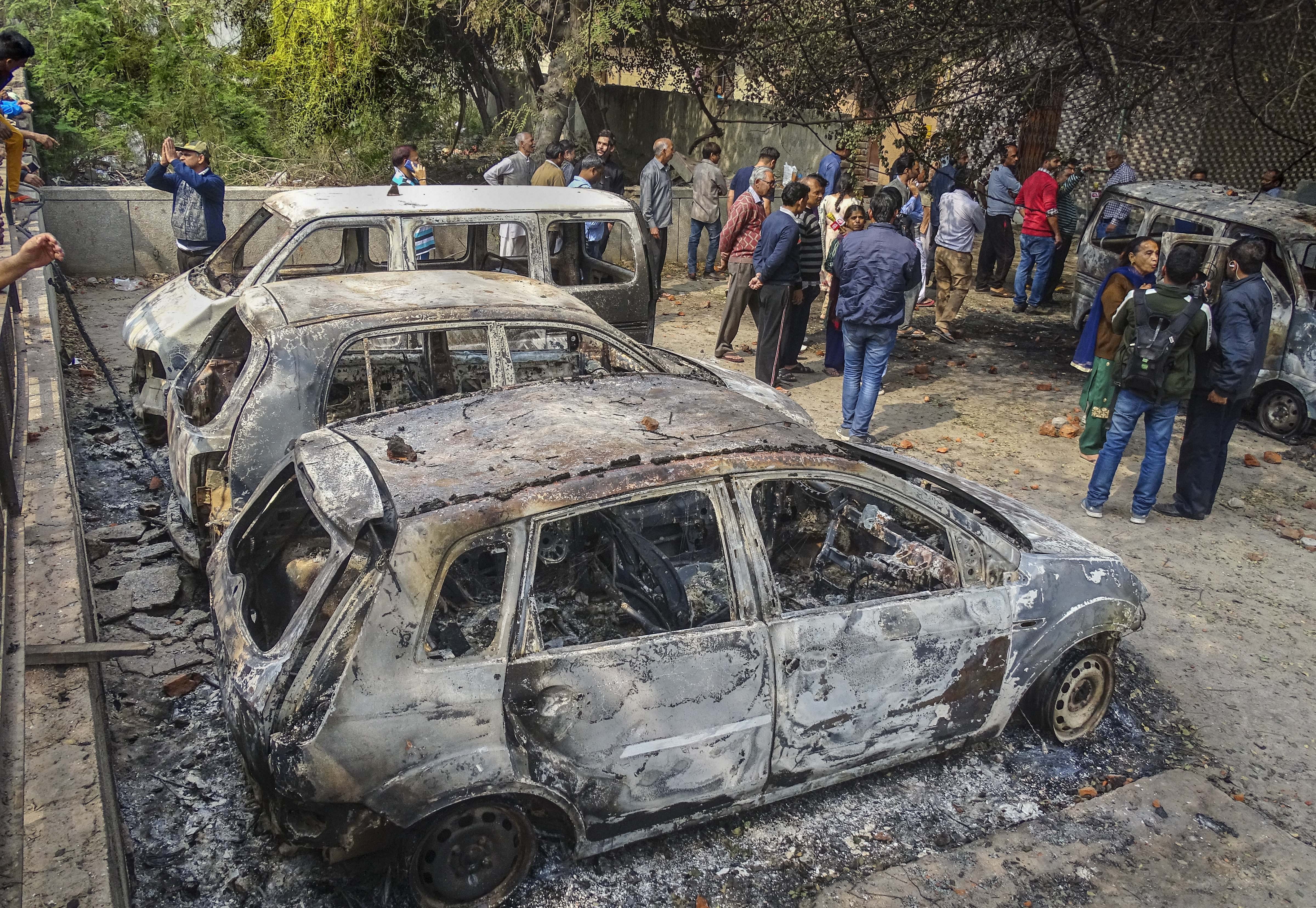 Passersby stand near vehicles which were set ablaze by rioters following clashes over the new citizenship law, in Yamuna Vihar area of northeast Delhi, Wednesday, February 26, 2020. 