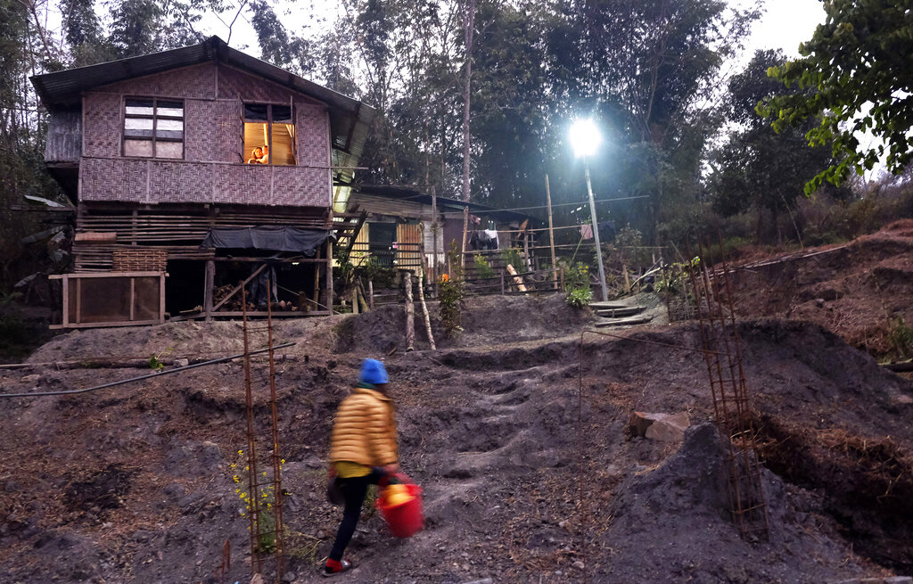 A young Naga girl walks with a bucket filled with pomelo collected from a nearby garden, as a woman holds a child and watches from the window in Kohima