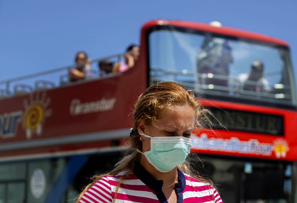 A woman wearing a mask as a precaution against the spread of the new coronavirus walks on the sidewalk as a tourist, double decker bus passes by in Havana, Cuba, Friday, March 20, 2020.