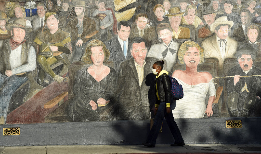 A woman wearing a mask passes in front of a mural depicting Hollywood legends as stay-at-home orders continue in California due to the coronavirus, Friday, April 3, 2020, in the Hollywood section of Los Angeles.