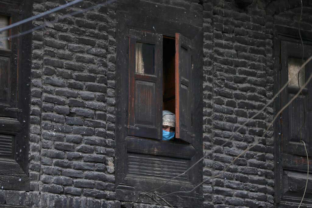In this April 6, 2020, file photo, a Kashmiri woman wearing protective mask watches through a window of her house as cops make markings for people to maintain social distance at a market in Srinagar.