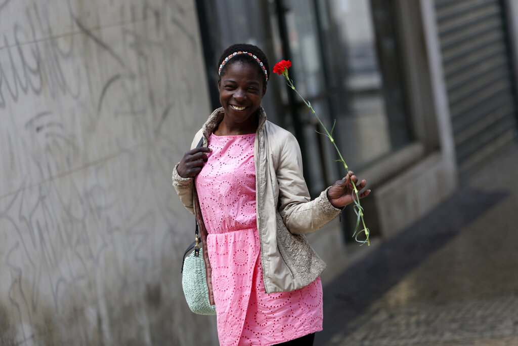 A young woman smiles while carrying a red carnation, symbol of the April 25th Portuguese revolution, in Lisbon's Mouraria neighborhood, Saturday, April 25, 2020. Portugal marks Saturday the anniversary of the 1974 revolution that restored democracy in the country, however due to the coronavirus outbreak this year without the celebrations that usually attract hundreds of thousands.