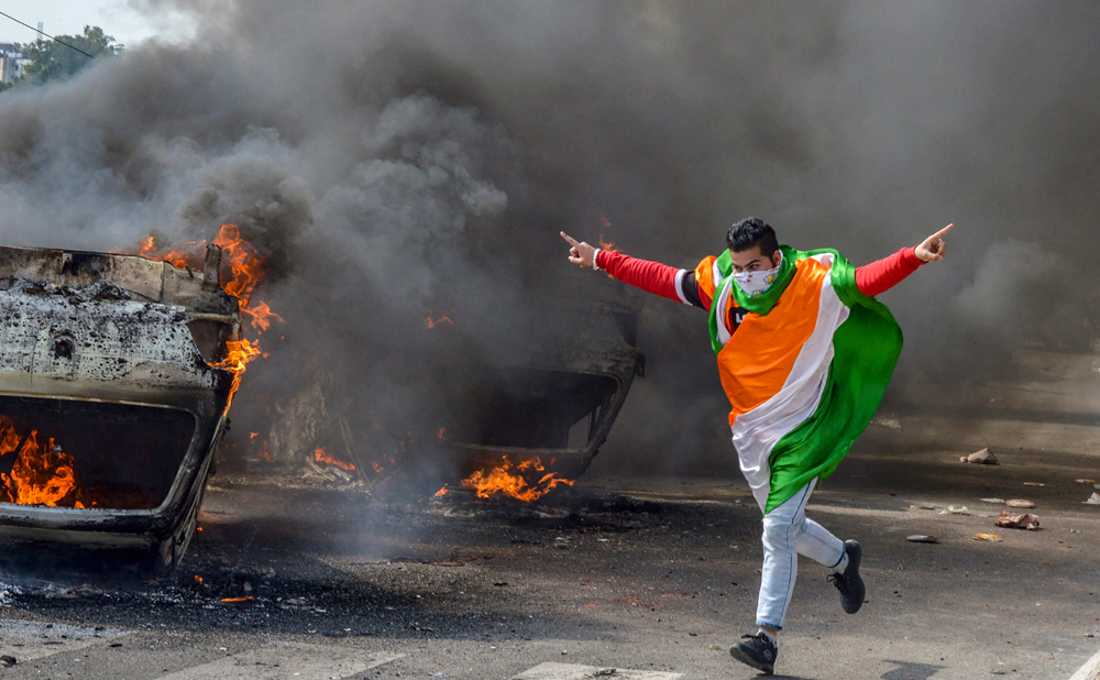 A protester draped in the Tricolour runs past burning vehicles that were set on fire during a protest against Thursday's Pulwama terror attack, in Jammu, on Friday, February 15, 2019. 